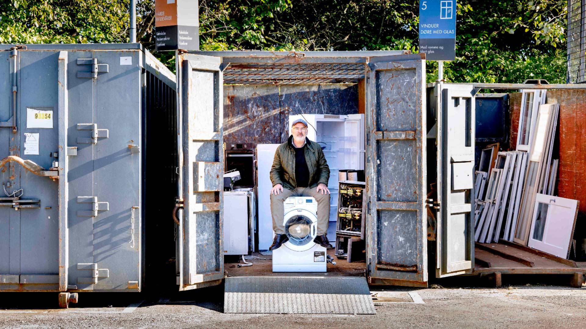 mand sidder på en kasseret vaskemaskine i en affaldscontainer 