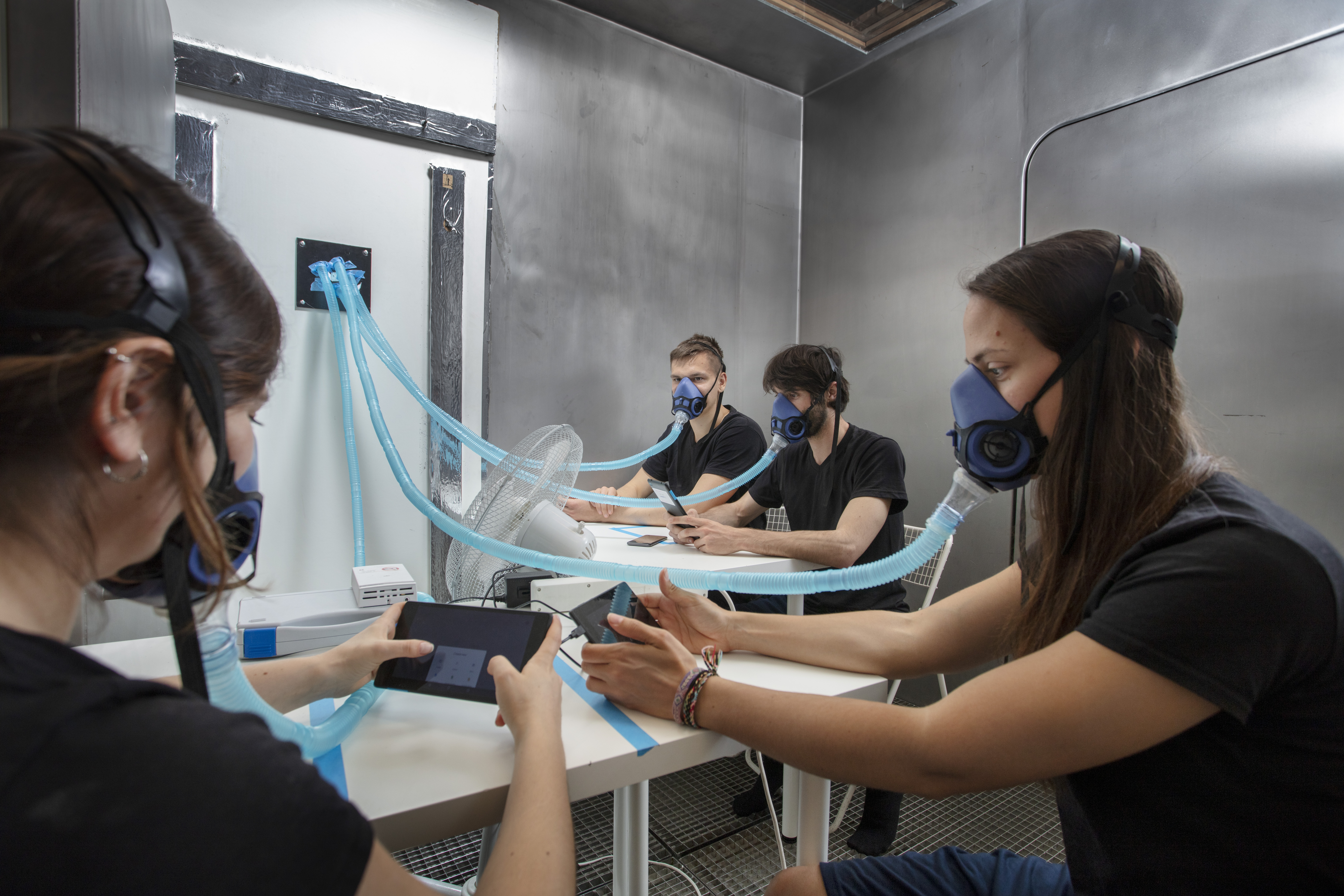 Four people wearing breathing masks while viewing a small tabled device in a sealed chamber